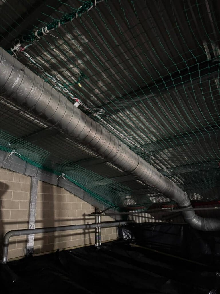 A large metal ventilation duct runs diagonally across an industrial ceiling, which is covered with green safety netting. The wall is made of beige bricks, and pipes are visible along the lower part of the image.
