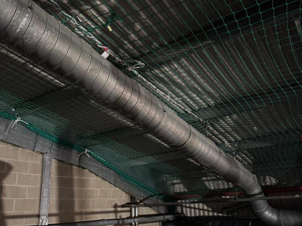 A large metal ventilation duct runs along the ceiling of an industrial building, with green safety netting installed above and along the walls. The walls are made of concrete blocks.