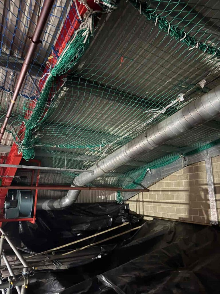 Industrial ceiling with exposed pipes, large ductwork, green safety nets, red metal framework, and a brick wall in the background. Black plastic sheeting hangs below, possibly for protection or ongoing construction.