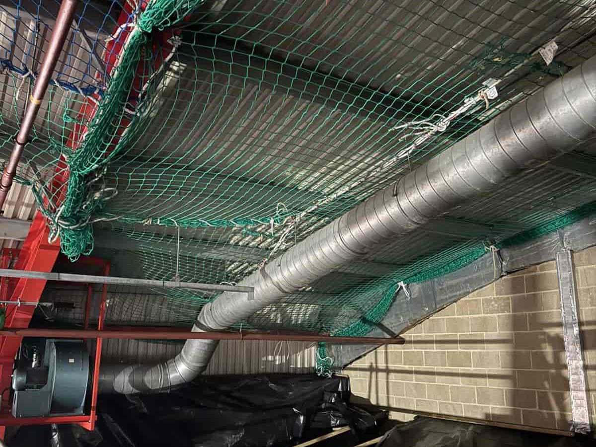 A silver ventilation duct runs diagonally across a ceiling lined with green safety netting. Red metal framework and pipes are visible on the left, while light brown brick walls appear on the right.