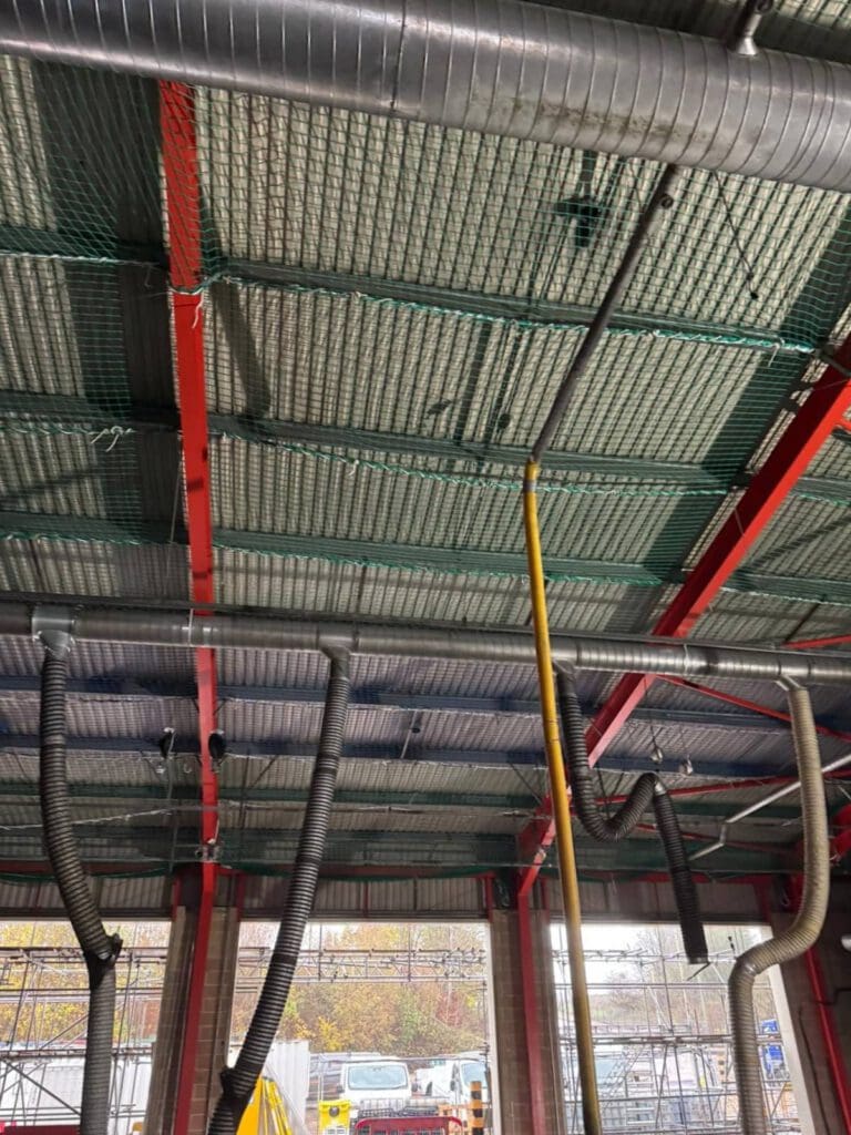 Industrial building interior showing exposed red steel beams, silver insulation, green safety netting, and hanging ventilation hoses. Outside, scaffolding and a white vehicle are visible through open walls.