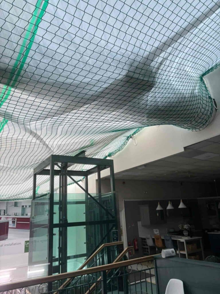 A large green net is suspended from the ceiling of an indoor space, bulging downward and collecting water from a leak above. Nearby are glass railings, part of a staircase, and a glass elevator shaft.