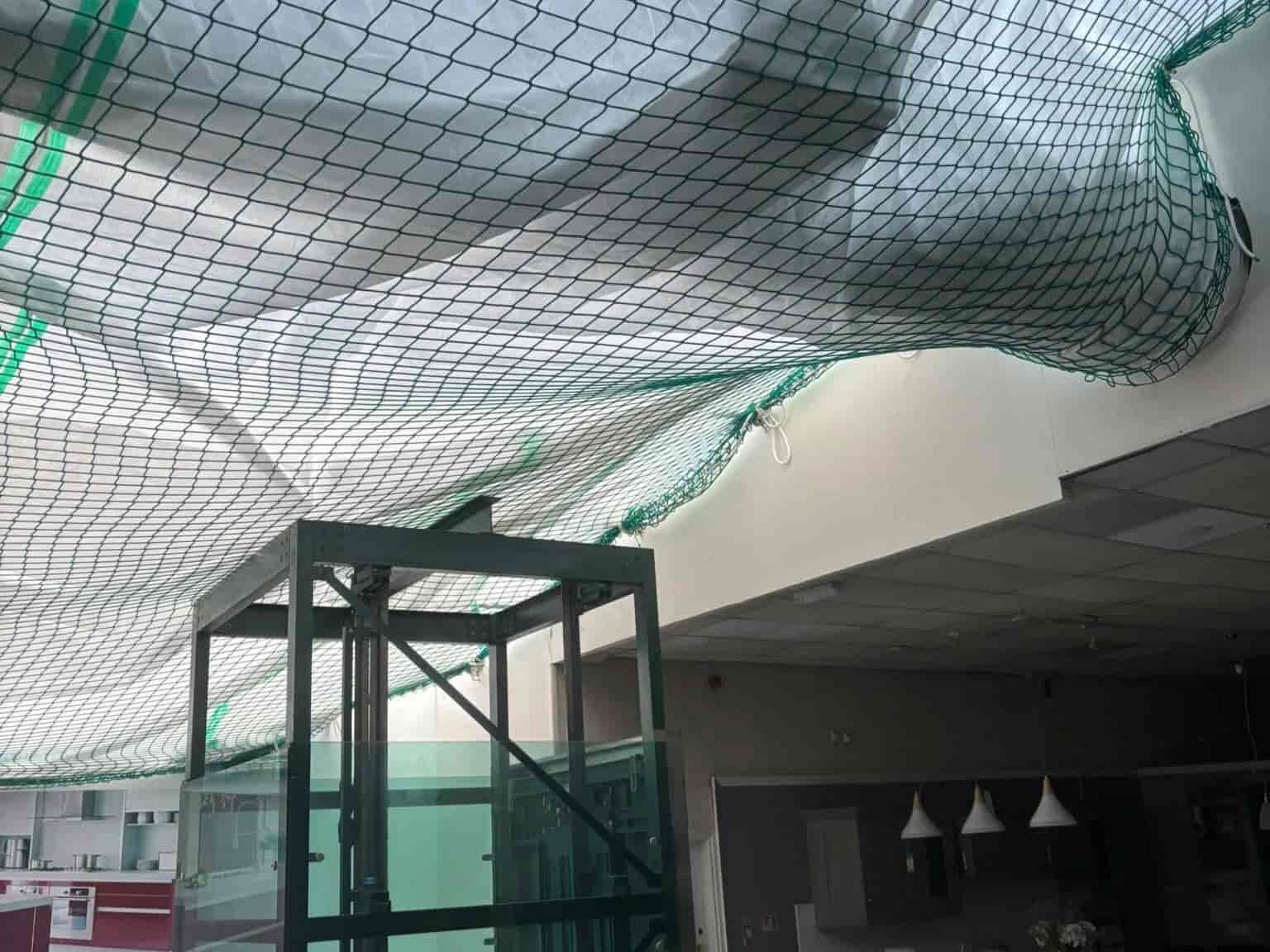 A green safety net is suspended across the ceiling of an indoor space, possibly under construction or repair. Below, there is a glass elevator shaft and some modern hanging lights.