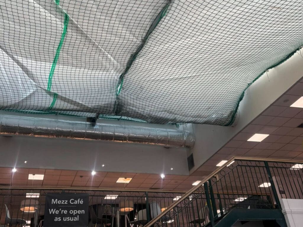 A café interior with ceiling covered by plastic sheets and netting, possibly for repairs. Metal ducts and ceiling lights are visible. A sign reads, "Mezz Café We’re open as usual." Stairs with a green railing are at the right.