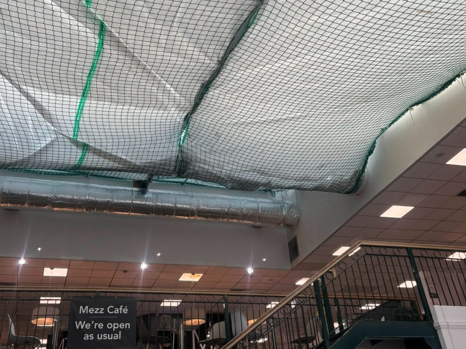 A café interior with ceiling covered by plastic sheets and netting, possibly for repairs. Metal ducts and ceiling lights are visible. A sign reads, "Mezz Café We’re open as usual." Stairs with a green railing are at the right.